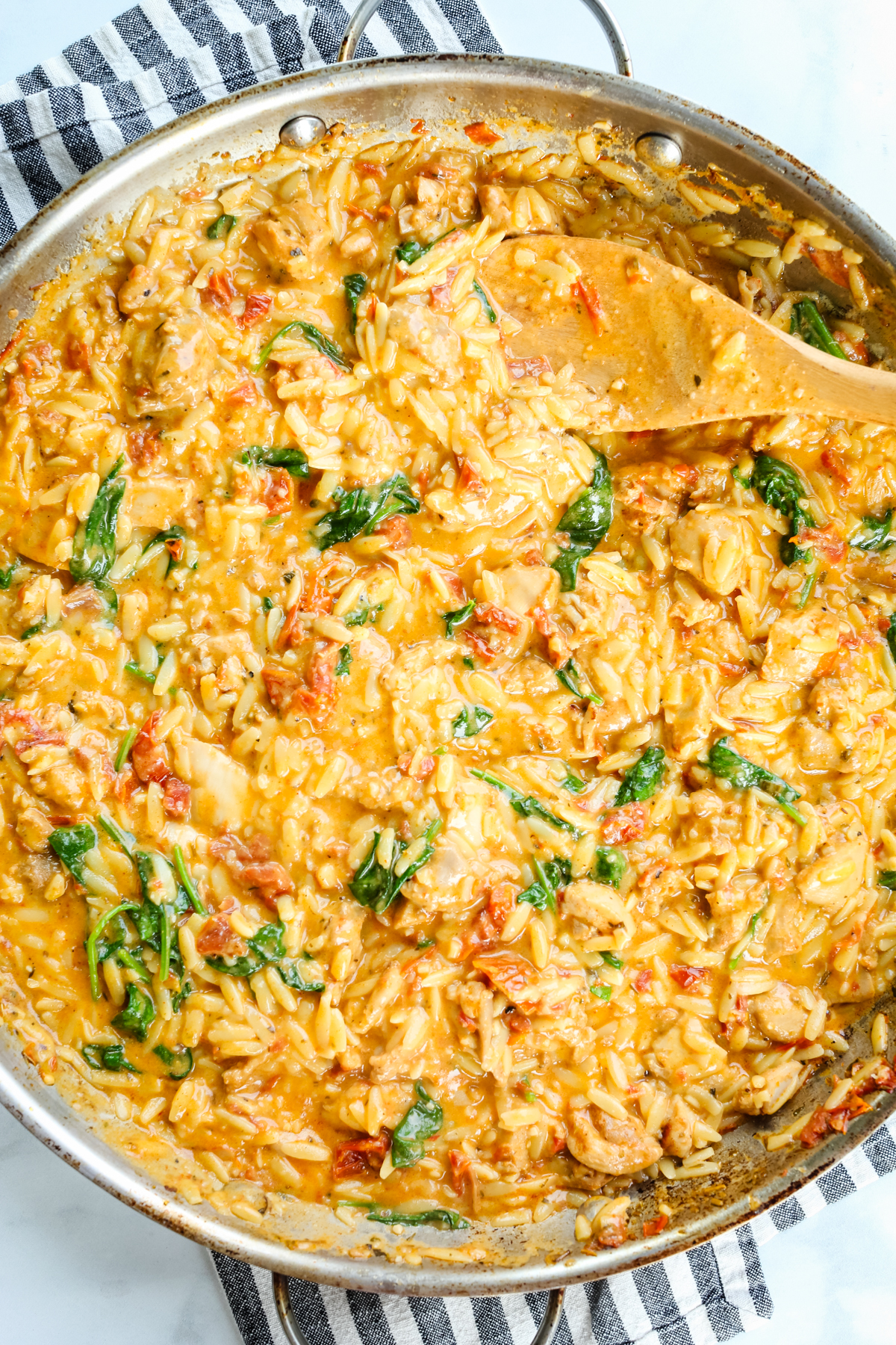 the skillet pan of marry me chicken orzo with a wooden spoon and a striped napkin under the pan