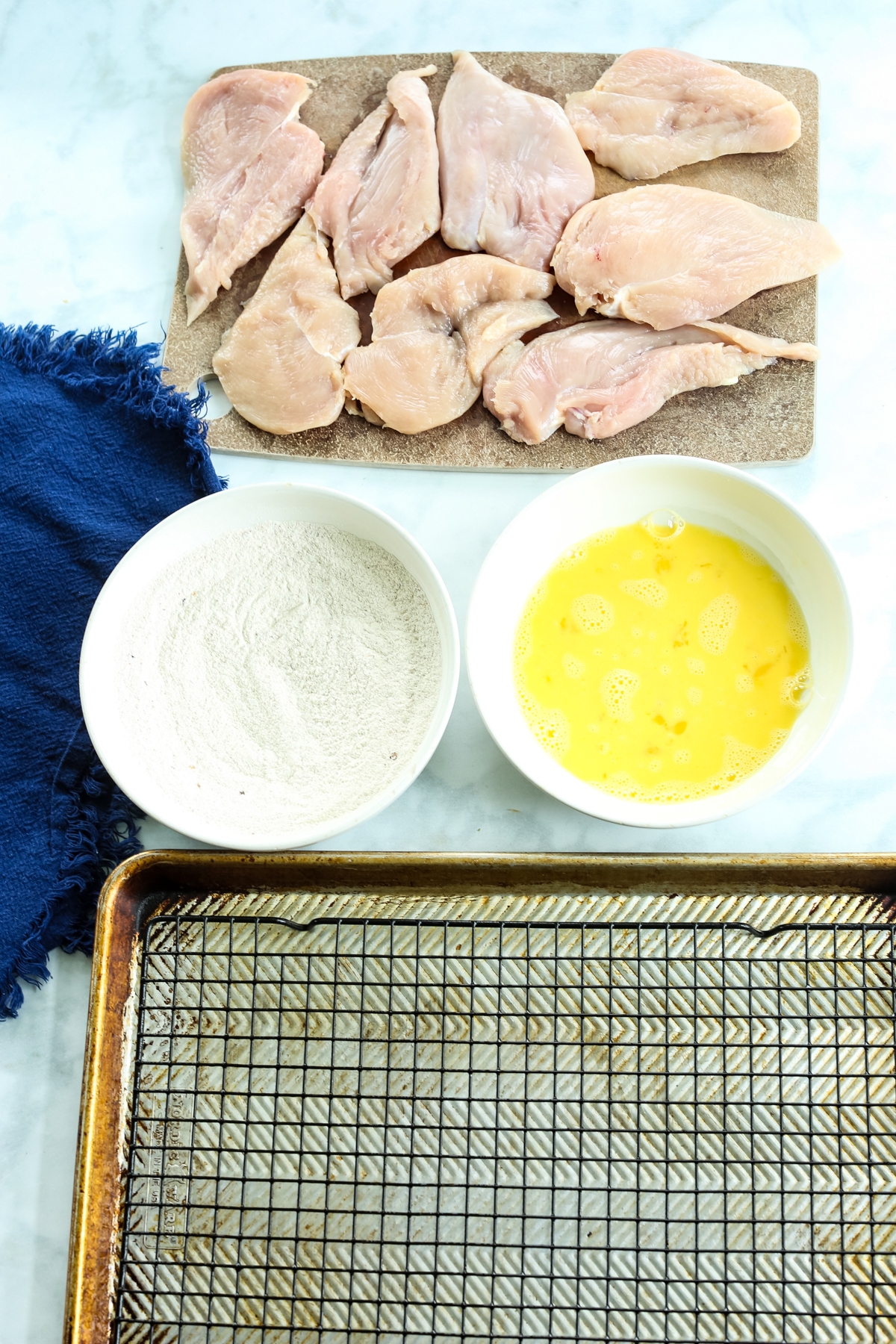 a line up of chicken, egg, flour, and a baking sheet with a wire rack on it. 