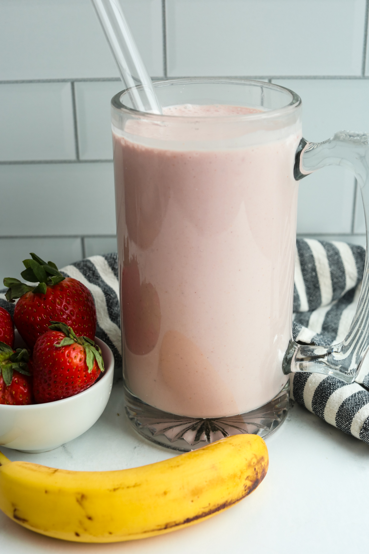 a large mug of strawberry banana protein smoothie with a small bowl of fresh strawberries and an unpeeled banana